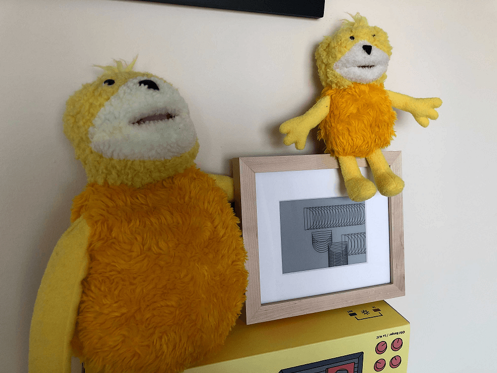 A photo of two stuffed animals next to a wooden frame with a digital screen in the middle of it. The screen contains a piece of art consisting of semicircles stacked on top of each other tightly, almost resembling a Slinky, scattered around the canvas